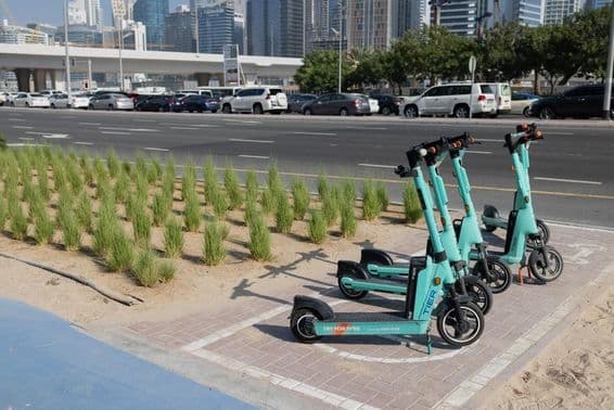 E-kolobežky zaparkované pred dubajským mrakodrapom s zelenými rastlinami na cestnom deliacom páse.