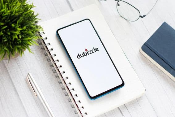 Mobil s aplikáciou Dubizzle na stole, pod poznámkovým blokom, vedľa okuliarov a malej zelenej rastliny.