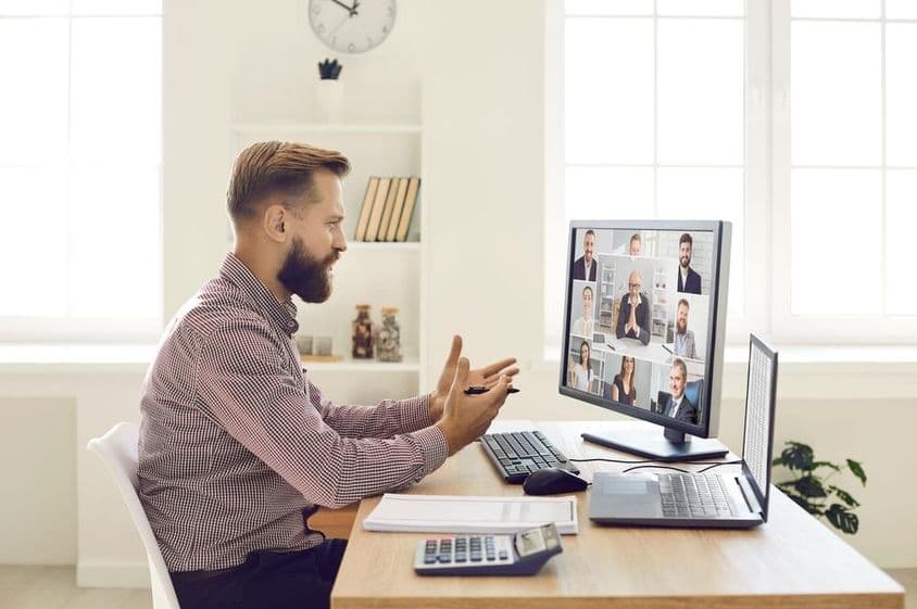 Osoba v kancelárskom prostredí s poznámkovým blokom a kalkulačkou na stole, vedúca videokonferenciu na monitore so zabudovanou webkamerou.
