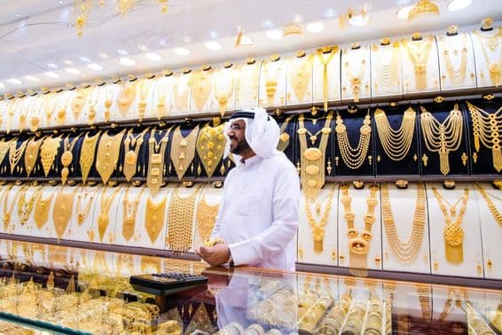 Dubajský predajca zlata s pozadím mnohých zlatých náhrdelníkov, predajca v tradičnom bielom oblečení.