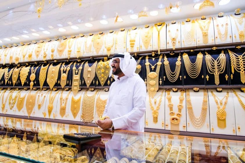 Dubajský predajca zlata s pozadím mnohých zlatých náhrdelníkov, predajca v tradičnom bielom oblečení.