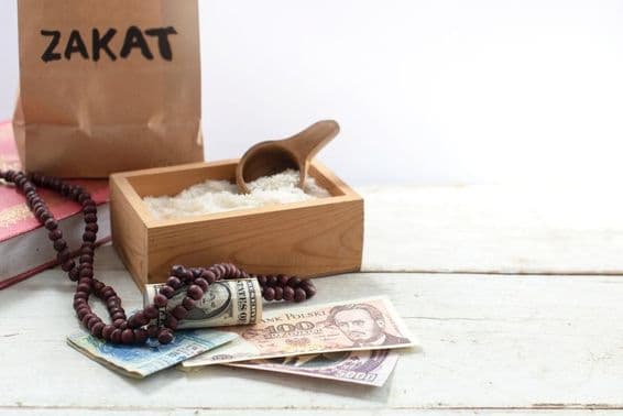 Taška označená zakat pred krabicou s ryžou, perlovými korálkami, dolármi a inými menami.