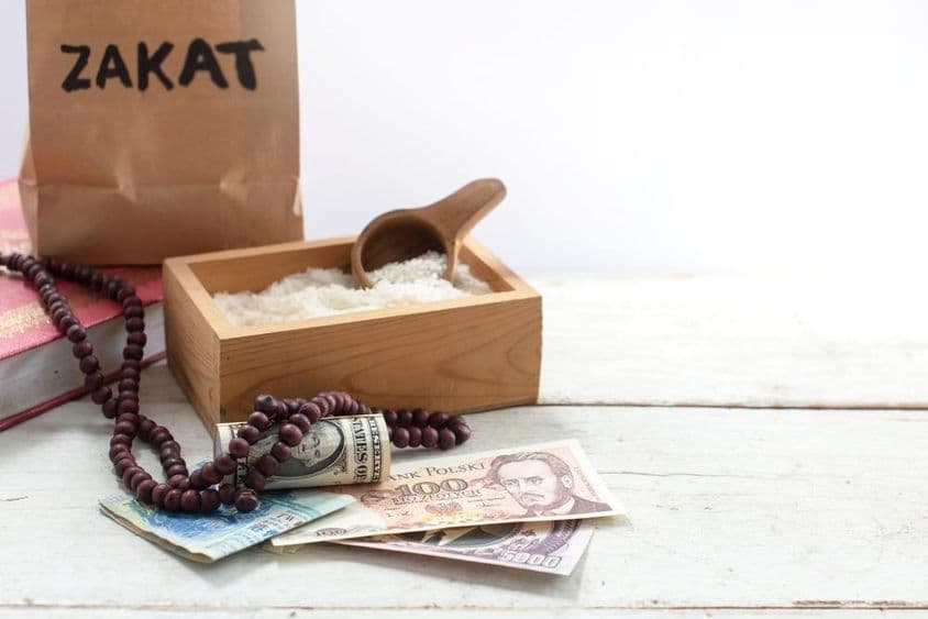 Taška označená zakat pred krabicou s ryžou, perlovými korálkami, dolármi a inými menami.
