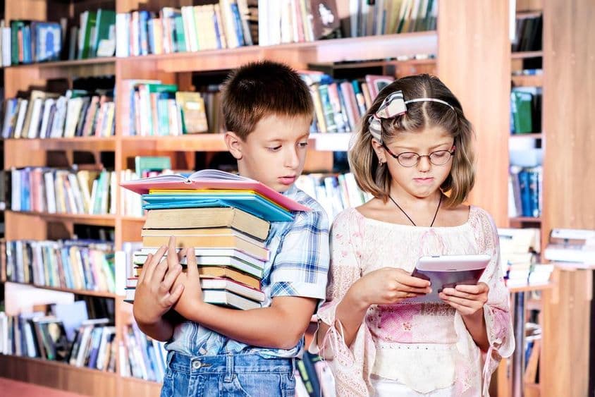 Dve deti v knižnici, jedno číta e-knihu, druhé nesie veľa kníh v rukách.