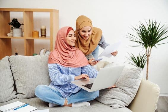 Dve emiratské ženy kontrolujúce zmluvu a notebook, jedna držiaca telefón, sediace na pohovke.