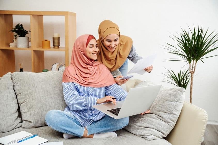 Dve emiratské ženy kontrolujúce zmluvu a notebook, jedna držiaca telefón, sediace na pohovke.