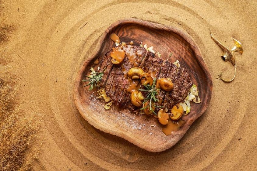Steak servírovaný s hubami na drevenom tanieri v púštnom piesku, so zhasnutou lampou vedľa.