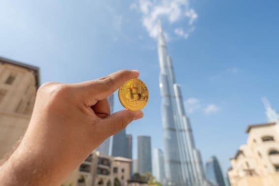 Osoba drží zlatú Bitcoin mincu, pozerajúca na oblohu, s Burdž Chalífou v pozadí.