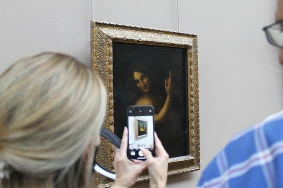 Obraz Svätého Jána Krstiteľa na stene, s ženou fotografujúcou ho telefónom.