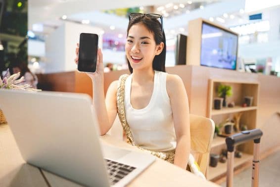 Dievča z generácie Z pracuje, drží telefón, pred ňou je notebook a vedľa nej rukoväť rolovacieho kufra.