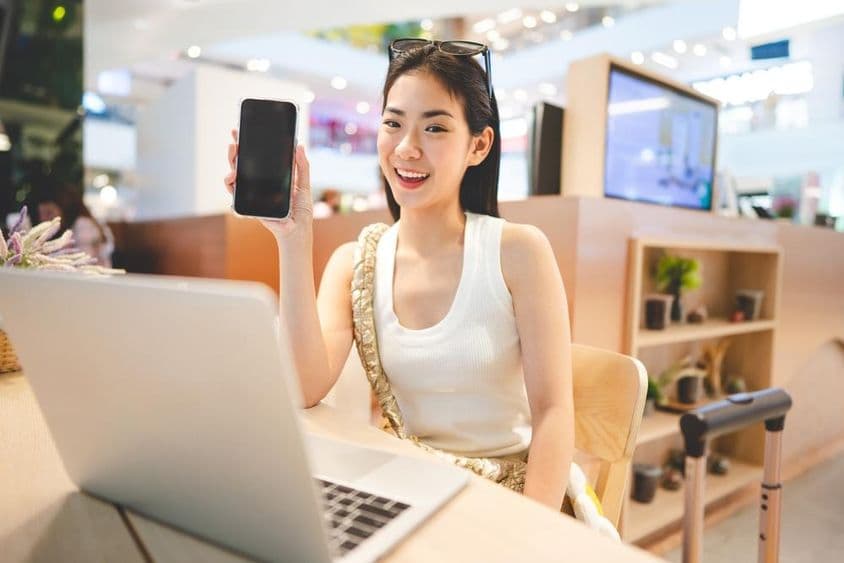 Dievča z generácie Z pracuje, drží telefón, pred ňou je notebook a vedľa nej rukoväť rolovacieho kufra.
