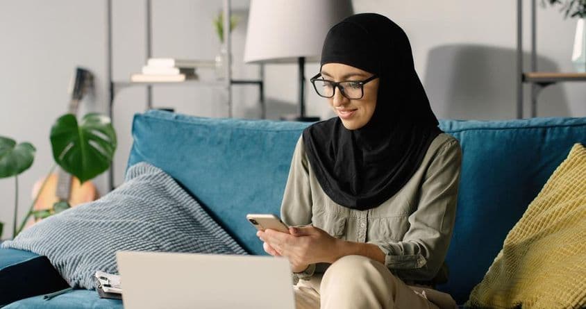 Arabská dievčina v hidžábe pracuje doma pomocou smartfónu a notebooku, sedia na pohovke.