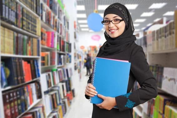 Arabský študent stojí v knižnici s notebookom v ruke.