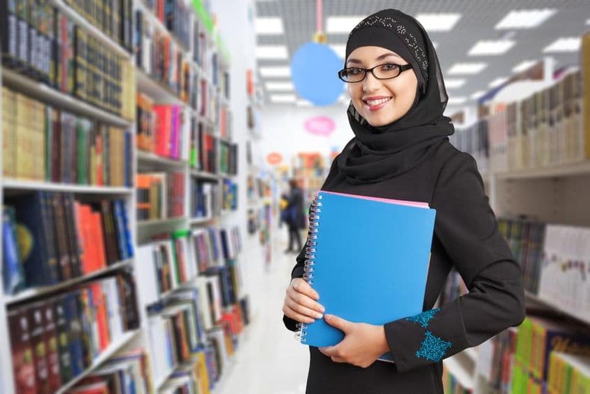 Arabský študent stojí v knižnici s notebookom v ruke.