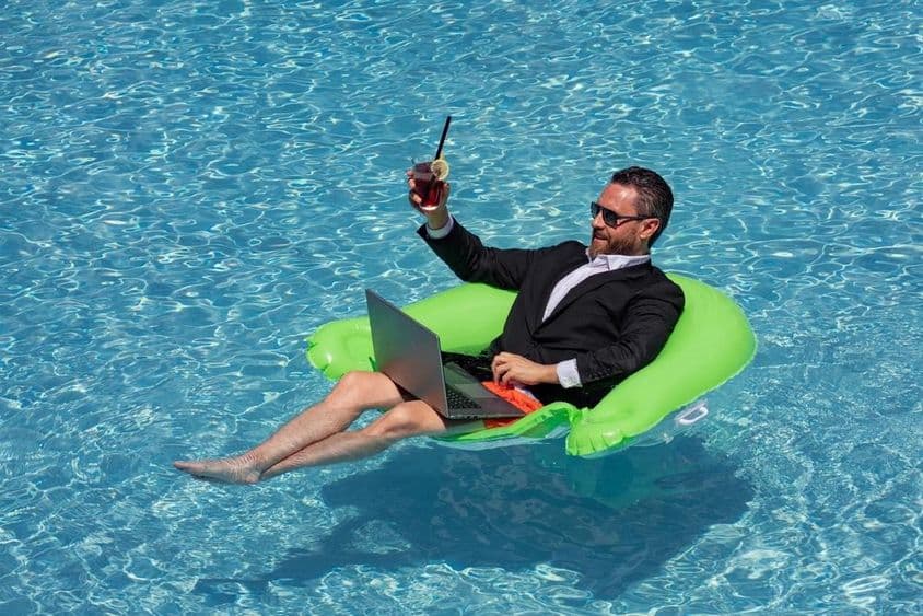 Úspešný podnikateľ v obleku plávajúci vo vode v bazéne, pijúci letný koktail a používajúci laptop v bazéne.