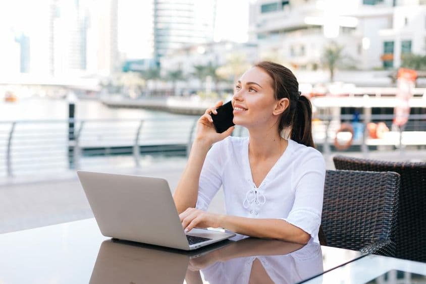 Mladá freelance žena pracuje na laptopu, sediaca v externej kaviarne držiac telefón.