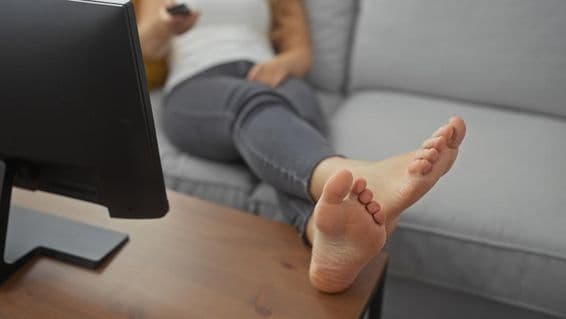 Mladá žena odpočívajúca naboso na útulnej pohovke v obývačke, držiaca diaľkový ovládač.