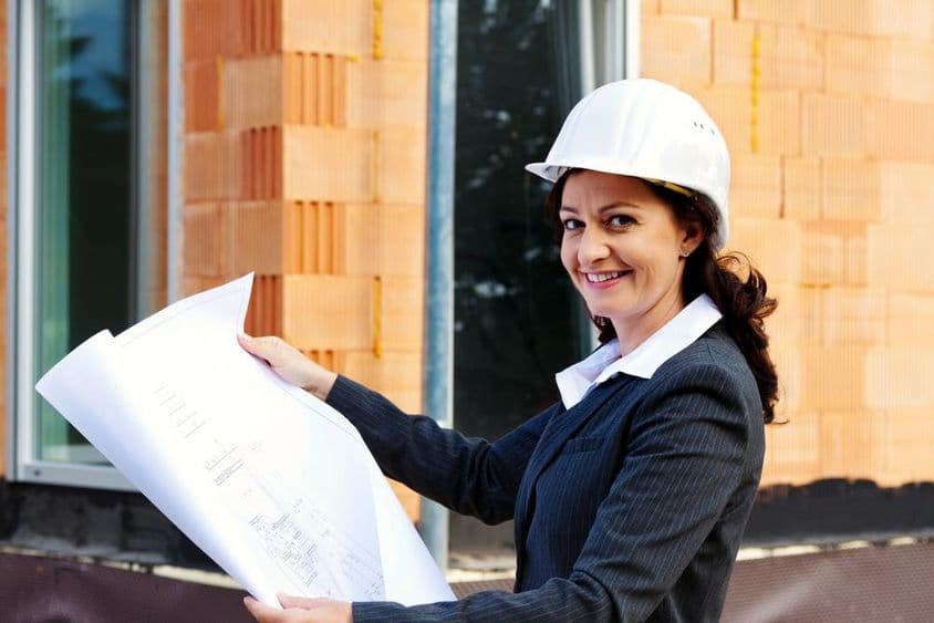 Žena architekta s plánom pred rozostavaným domom, nosiaca ochrannú prilbu.