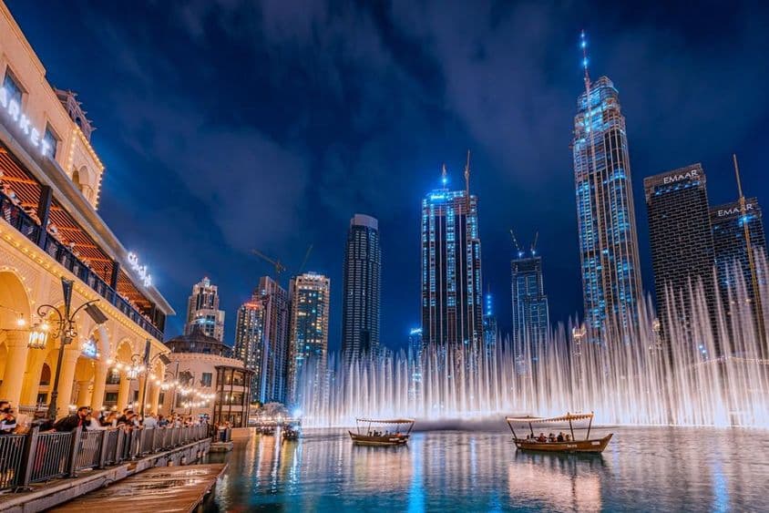 Ľudia na plavbe loďou sledujú Dubajskú fontánu v noci, so siluetou mrakodrapov v pozadí.