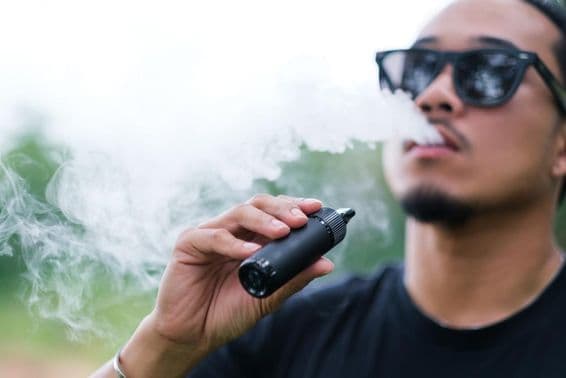 Ázijský muž fajčí s vapingovou elektronickou cigaretou, vyfukuje dym z úst a užíva si pobyt vonku v lete.