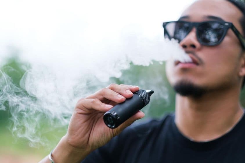 Ázijský muž fajčí s vapingovou elektronickou cigaretou, vyfukuje dym z úst a užíva si pobyt vonku v lete.