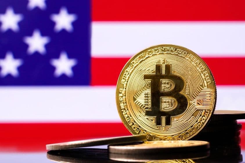 Zlatá kovová minca Bitcoinu s vlajkou USA v pozadí.