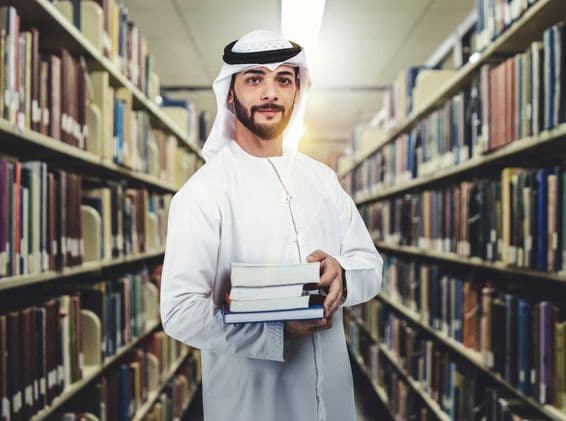Arabský študent stojaci v knižnici, držiaci knihy.