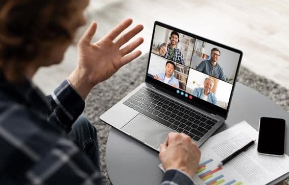 Muž sediaci pri stole, účastníci video konferencie na svojom notebooku.