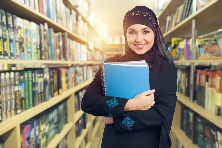 Arabský študent stojaci v knižnici s knihami v ruke.