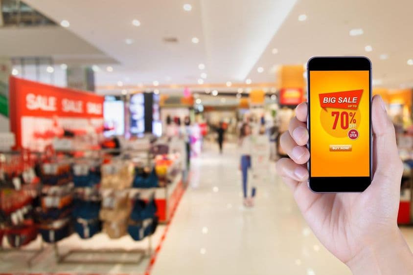 Smartfón so zobrazením aplikácie veľkého výpredaja na rozmazanom pozadí obchodného centra.