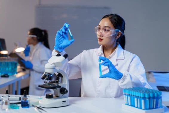 Ázijská vedkyňa v laboratórnom plášti a rukaviciach porovnáva dve testovacie skúmavky.