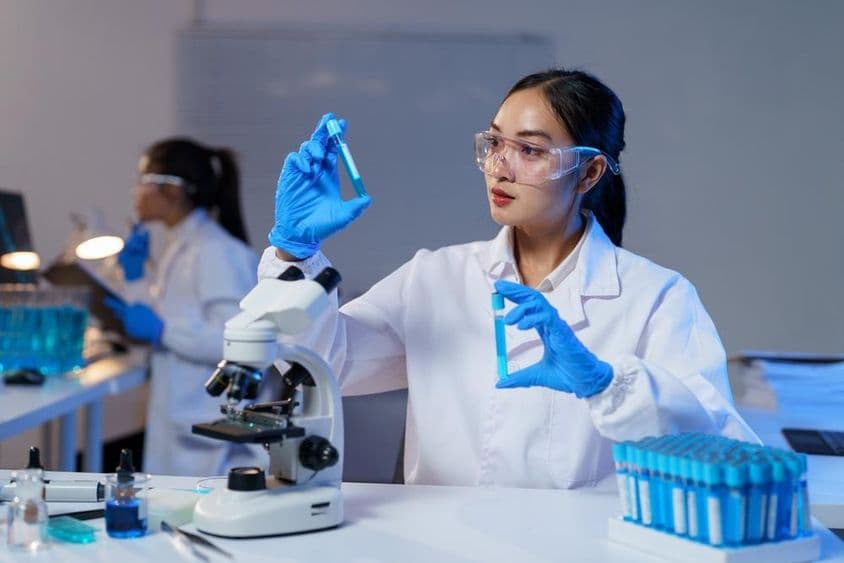 Ázijská vedkyňa v laboratórnom plášti a rukaviciach porovnáva dve testovacie skúmavky.