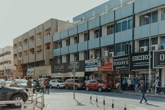 Rušná ulica v Bur Dubaji s klenotníctvami, chodcami a autami počas slnečného dňa.