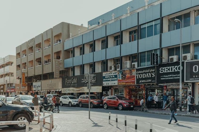 Rušná ulica v Bur Dubaji s klenotníctvami, chodcami a autami počas slnečného dňa.