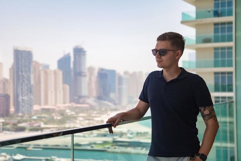 Mladý muž v okuliaroch stojaci na balkóne mrakodrapu s výhľadom na Dubaj.