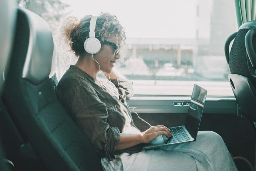 Mladá žena pracujúca na prenosnom počítači so slúchadlami na sedadle autobusu.
