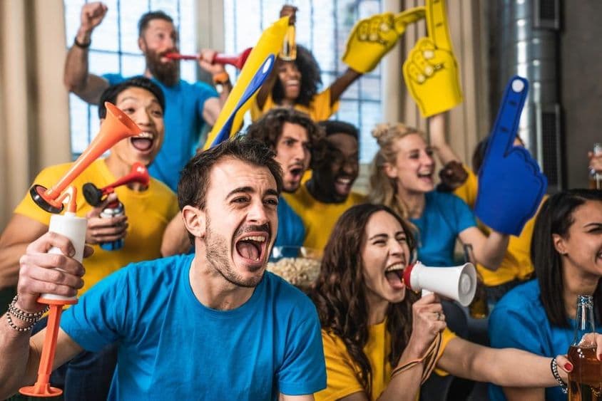 Národnostne rozmanití, radostní športoví fanúšikovia sledujúci zápas na televízii doma.