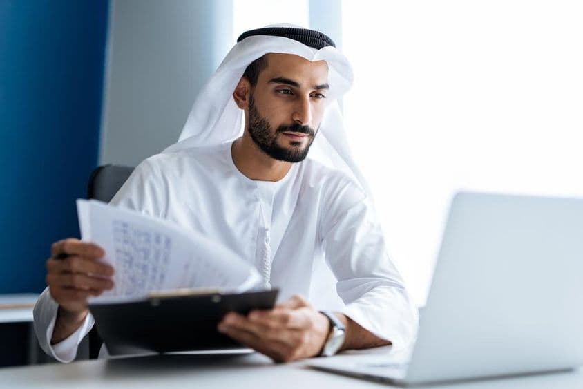 Muž pracujúci s laptopom v kancelárii v Dubaji, oblečený v tradičnom bielom emiratskom oblečení.