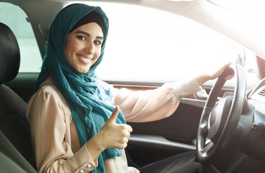 Šťastná arabská žena v hidžábe riadiaca auto so zdvihnutým palcom.