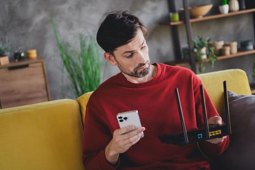 Mladý muž relaxuje doma so smartfónom a routerom, užíva si pokojnú popoludní.